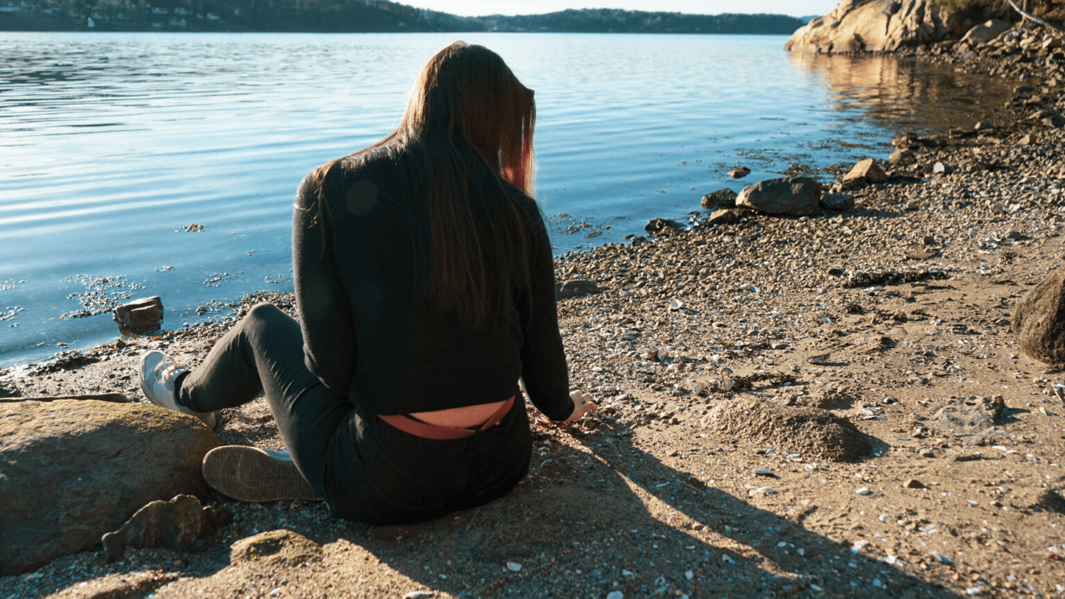 Person som sitter ved vannkanten med ryggen mot kameraet.