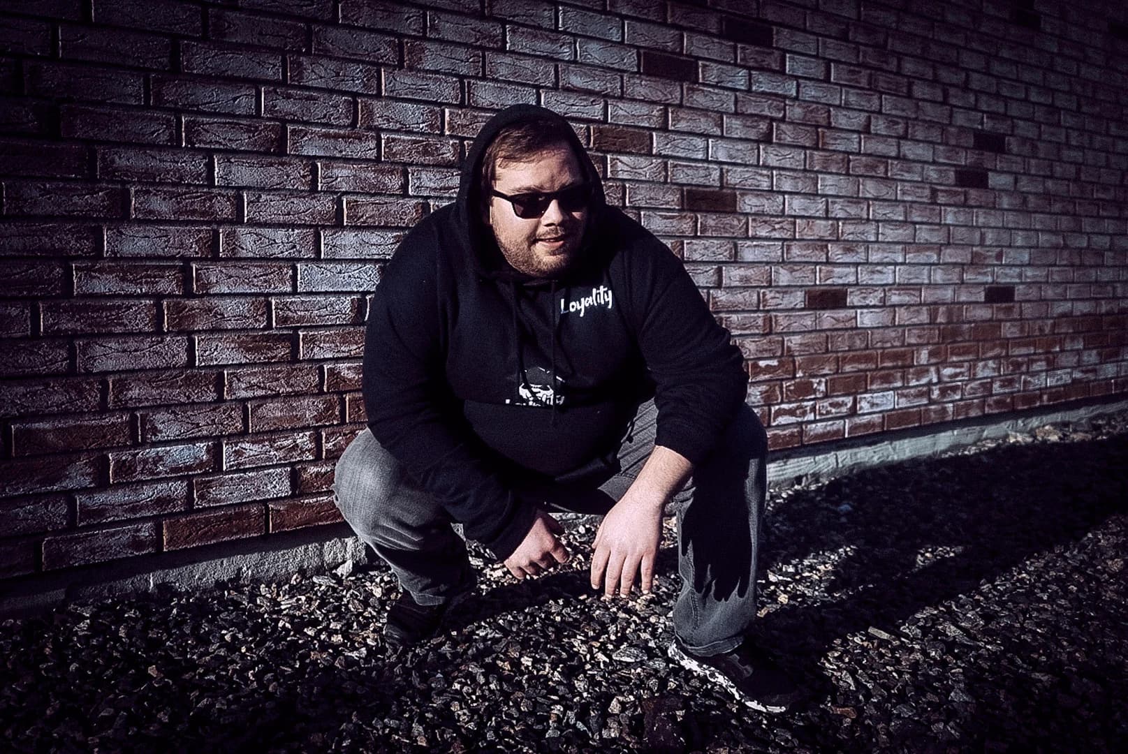 Mann med hettegenser og solbriller foran en mørk murvegg.